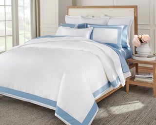 White and blue bedding set on a bed with a side table and flowers in the background.