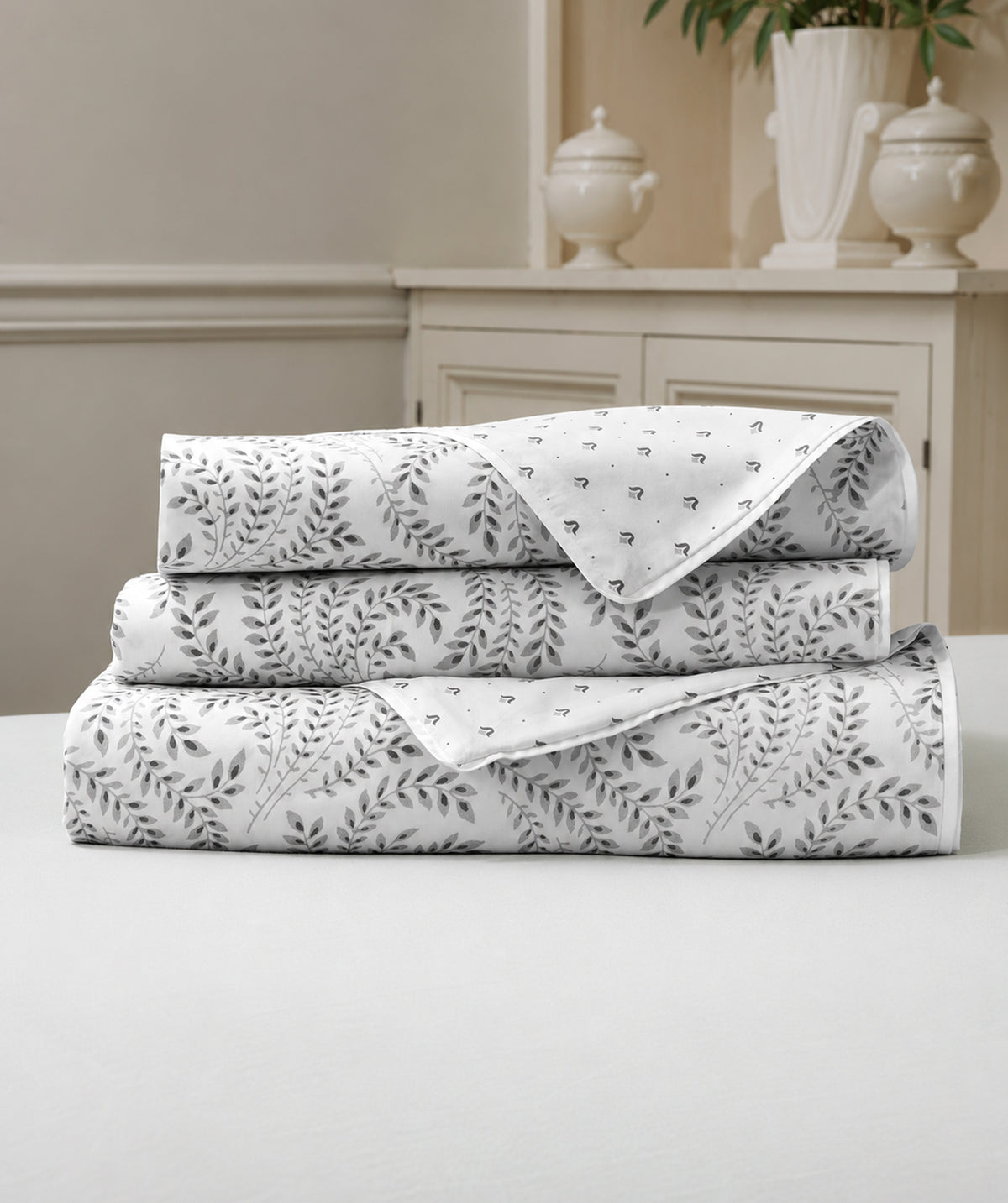 A stack of three folded white bedding sheets or linens with a gray botanical leaf pattern, displayed on a white table in front of a neutral beige interior background.