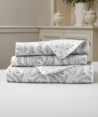 A stack of three folded white bedding sheets or linens with a gray botanical leaf pattern, displayed on a white table in front of a neutral beige interior background.