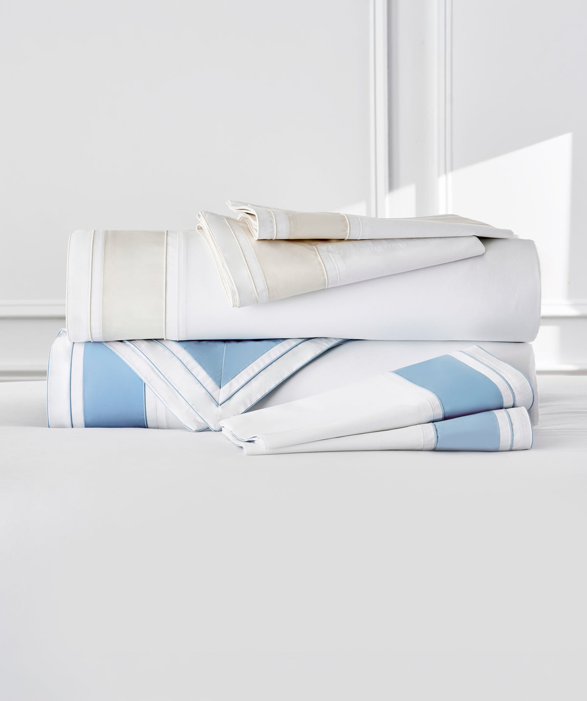 A stack of folded bedding linens in white, cream, and light blue with striped borders, arranged on a light gray surface against a bright white background.
