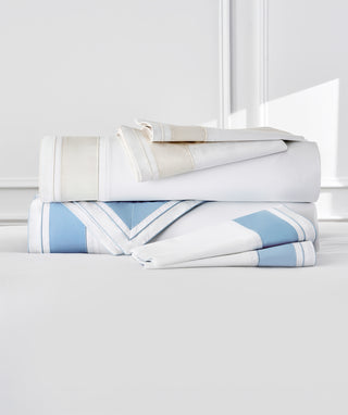 A stack of folded bedding linens in white, cream, and light blue with striped borders, arranged on a light gray surface against a bright white background.