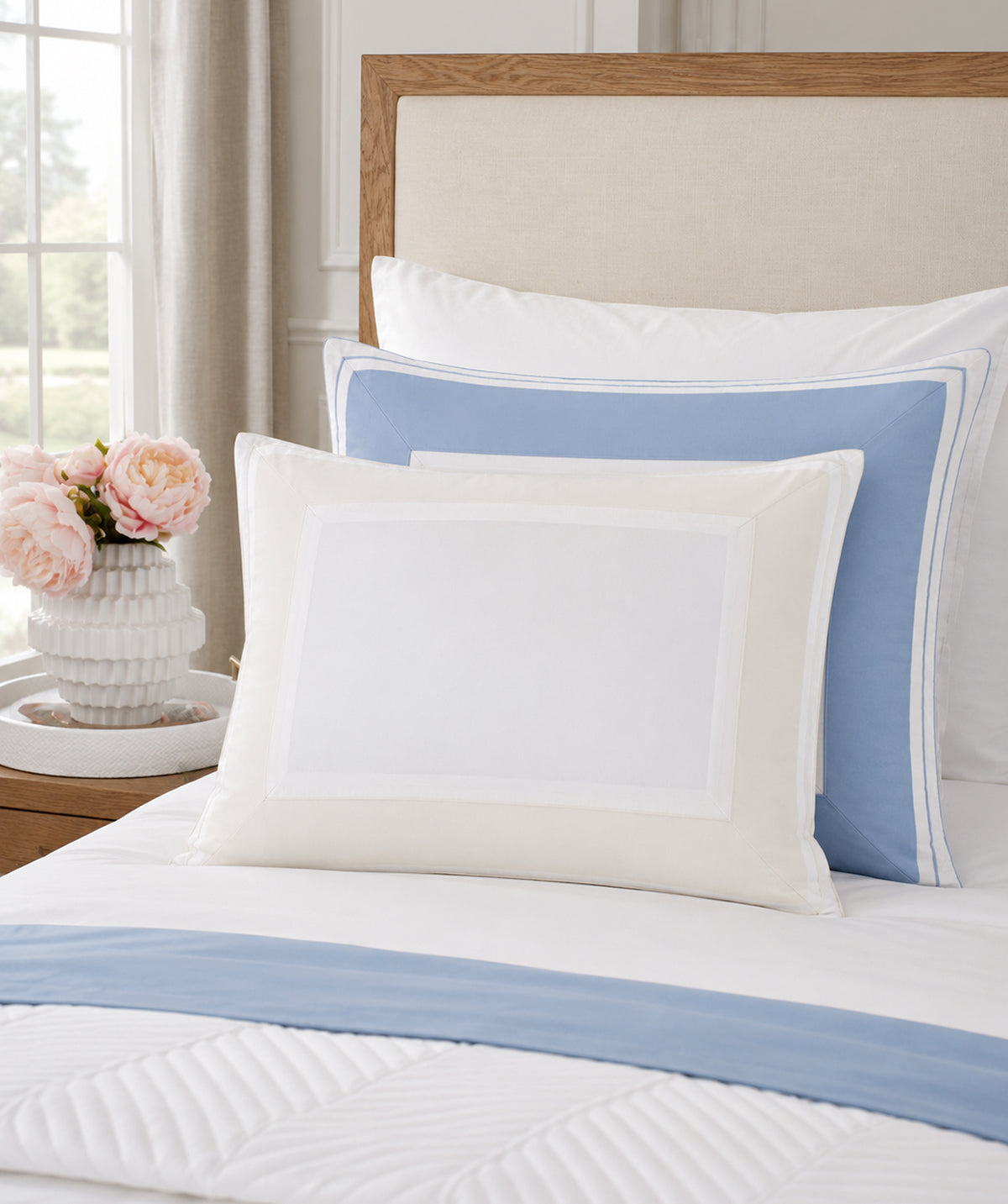 A styled bedroom scene showing layered bedding with white pillows, a light blue accent pillow, and a white quilted comforter with a blue stripe, set against a beige upholstered headboard. A small vase of pink flowers sits on a bedside table near a window.