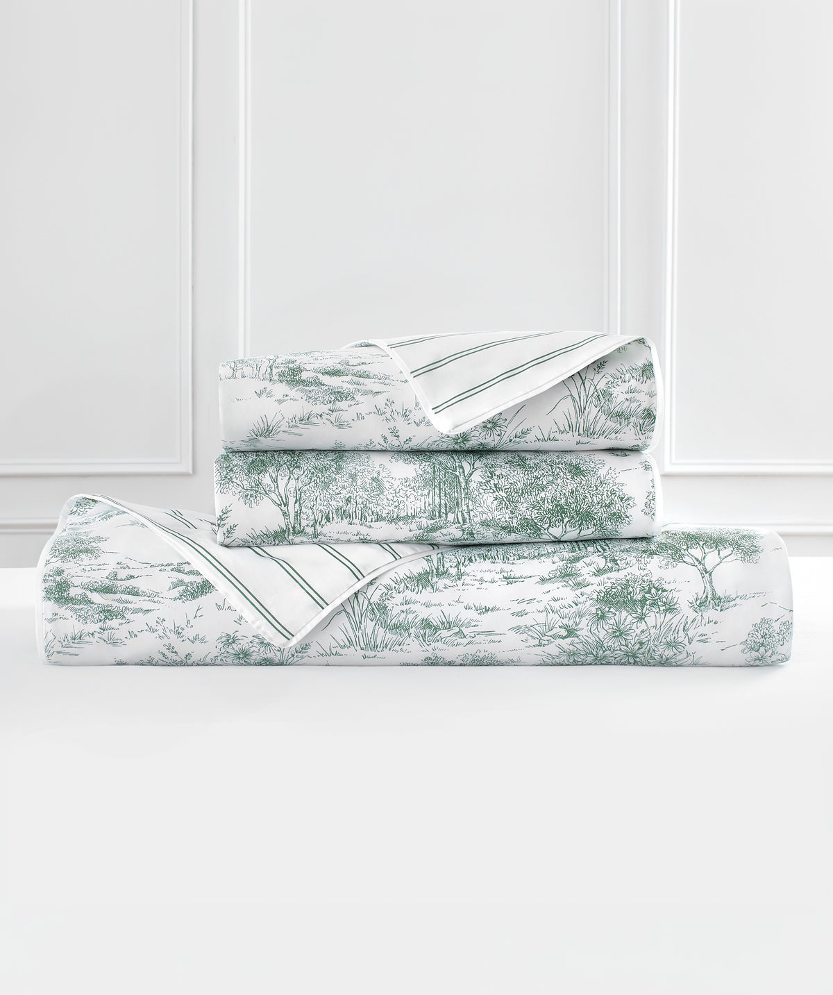 A stack of folded bedding, including a flat sheet and pillowcases, featuring a green toile de Jouy landscape pattern on a white background with thin green stripes.