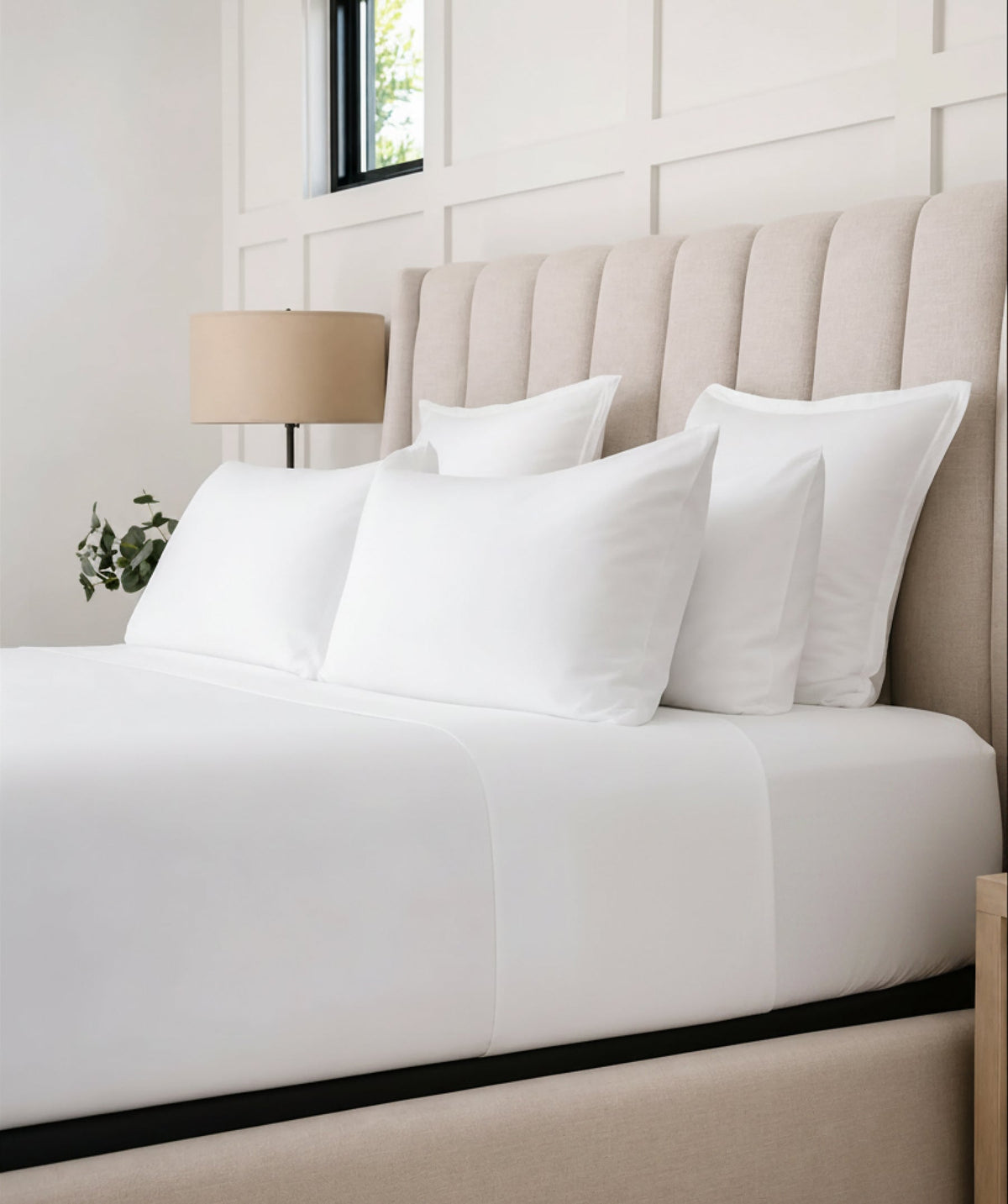 Bright white sheet set styled on a bed with layered pillows and a neutral upholstered headboard.