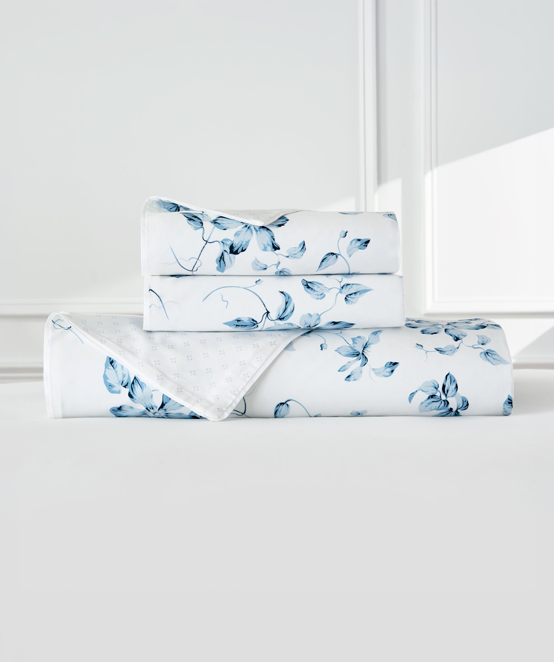 A folded bedding set with a blue floral print on a white background, stacked neatly on a white surface against a white paneled wall.