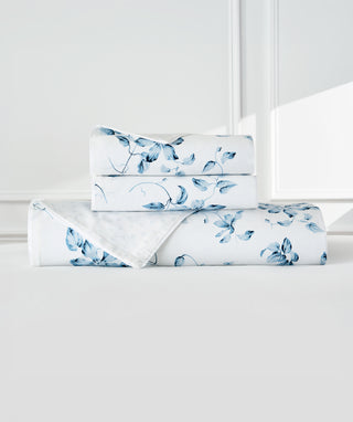 A folded bedding set with a blue floral print on a white background, stacked neatly on a white surface against a white paneled wall.
