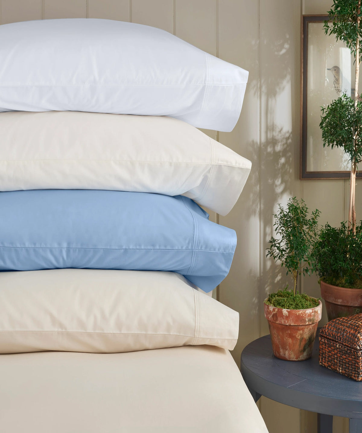 Stacked pillowcases in white, ivory, and powder blue styled beside potted greenery.