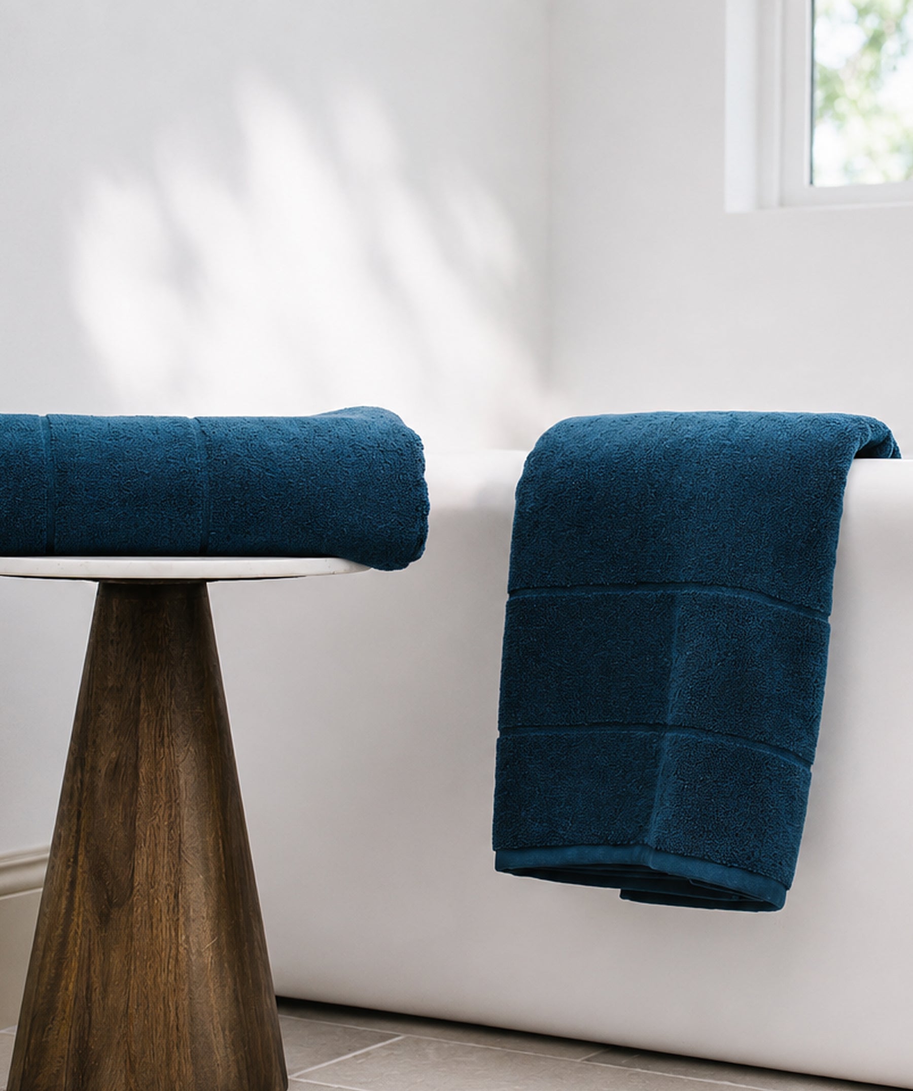 Navy bath sheet folded on a stool beside a white bathtub with a matching bath sheet draped over the tub.