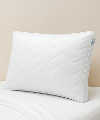 A white quilted pillow sham with a chevron pattern, resting on a white bed sheet against a beige wall.
