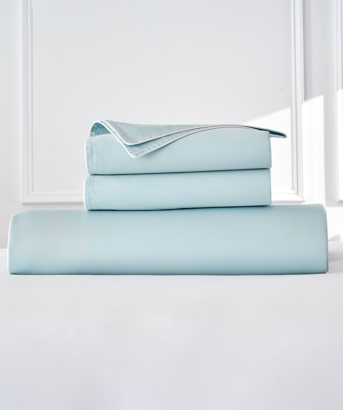 A stack of light blue bedding, including a duvet and two folded pillowcases, displayed on a white surface against a bright white background.