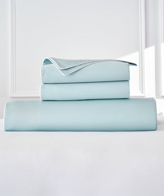 A stack of light blue bedding, including a duvet and two folded pillowcases, displayed on a white surface against a bright white background.