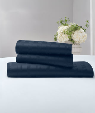 Three stacked dark navy blue textured sheets or linens on a white surface, with a vase of white flowers in the background against a light gray wall.